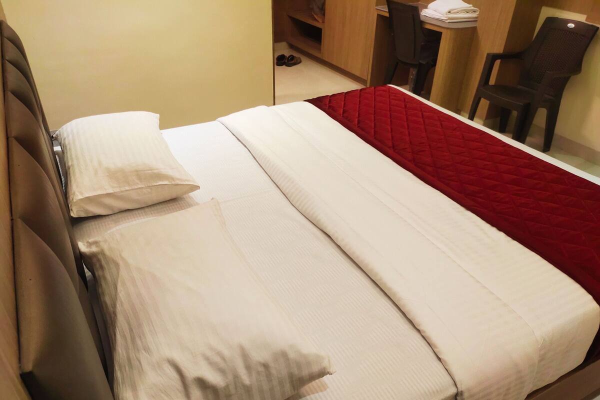 Una cama individual con ropa de cama blanca y un detalle rojo, un cabecero de madera, y una pequeña mesa con una silla al fondo.