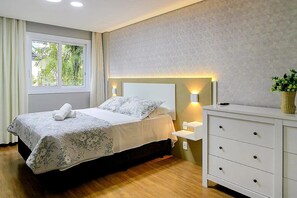 Quarto com cama, mesas de cabeceira, cômoda e janela com cortinas.