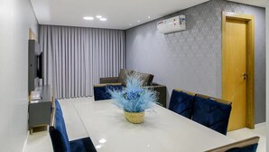 A modern dining area with a white table, blue chairs, a vase of blue flowers, a grey curtain, a patterned wall, and a wooden door. - Apto Novo a 400m da Borges de Medeiros (Gramado)