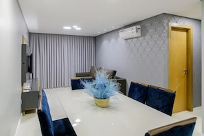 Uma zona de refeições moderna com uma mesa branca, cadeiras azuis, um vaso com flores azuis, uma cortina cinzenta, uma parede estampada e uma porta de madeira.