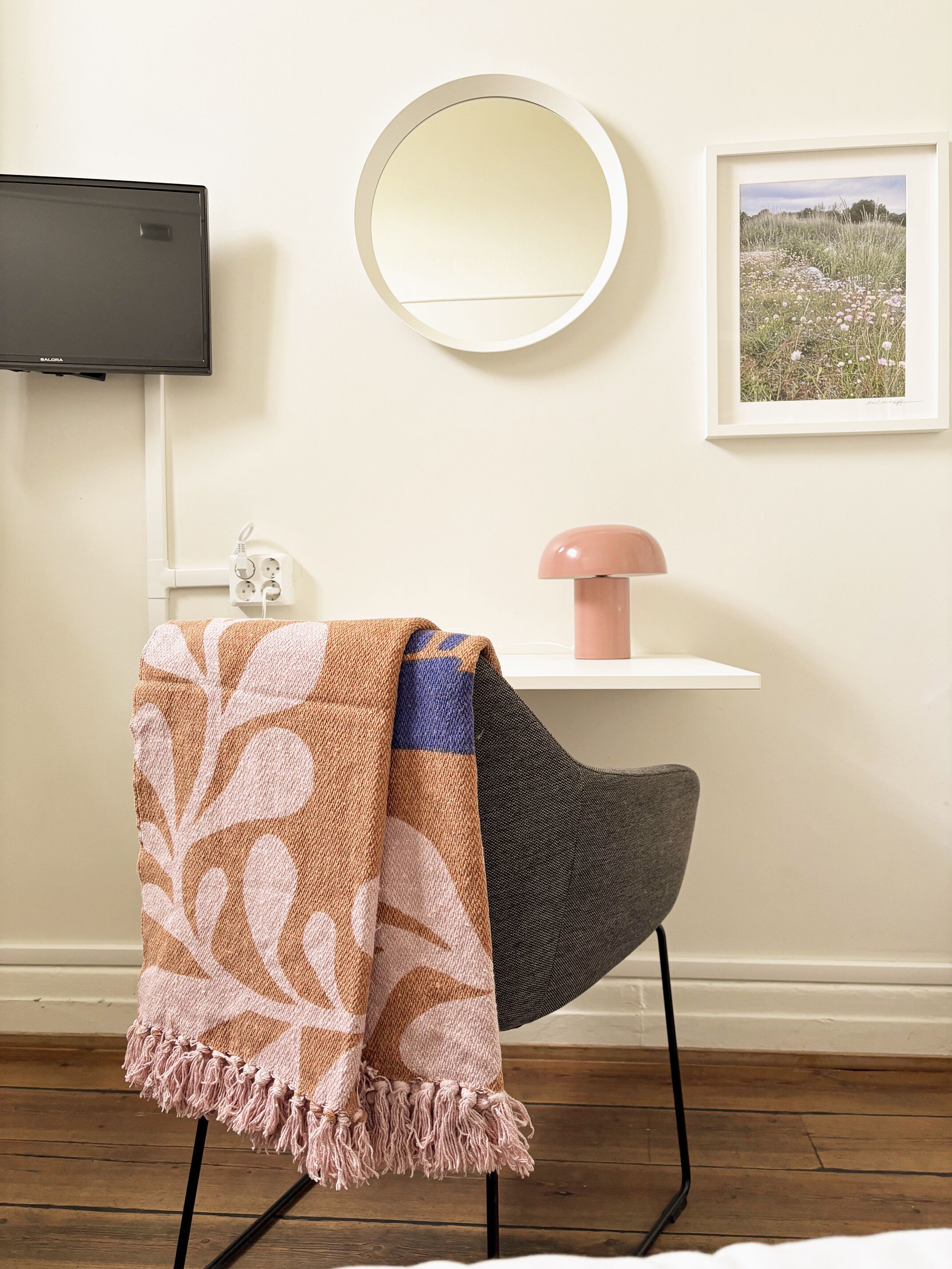 Una habitación con un espejo redondo, un cuadro enmarco, un televisor y una silla con una manta.