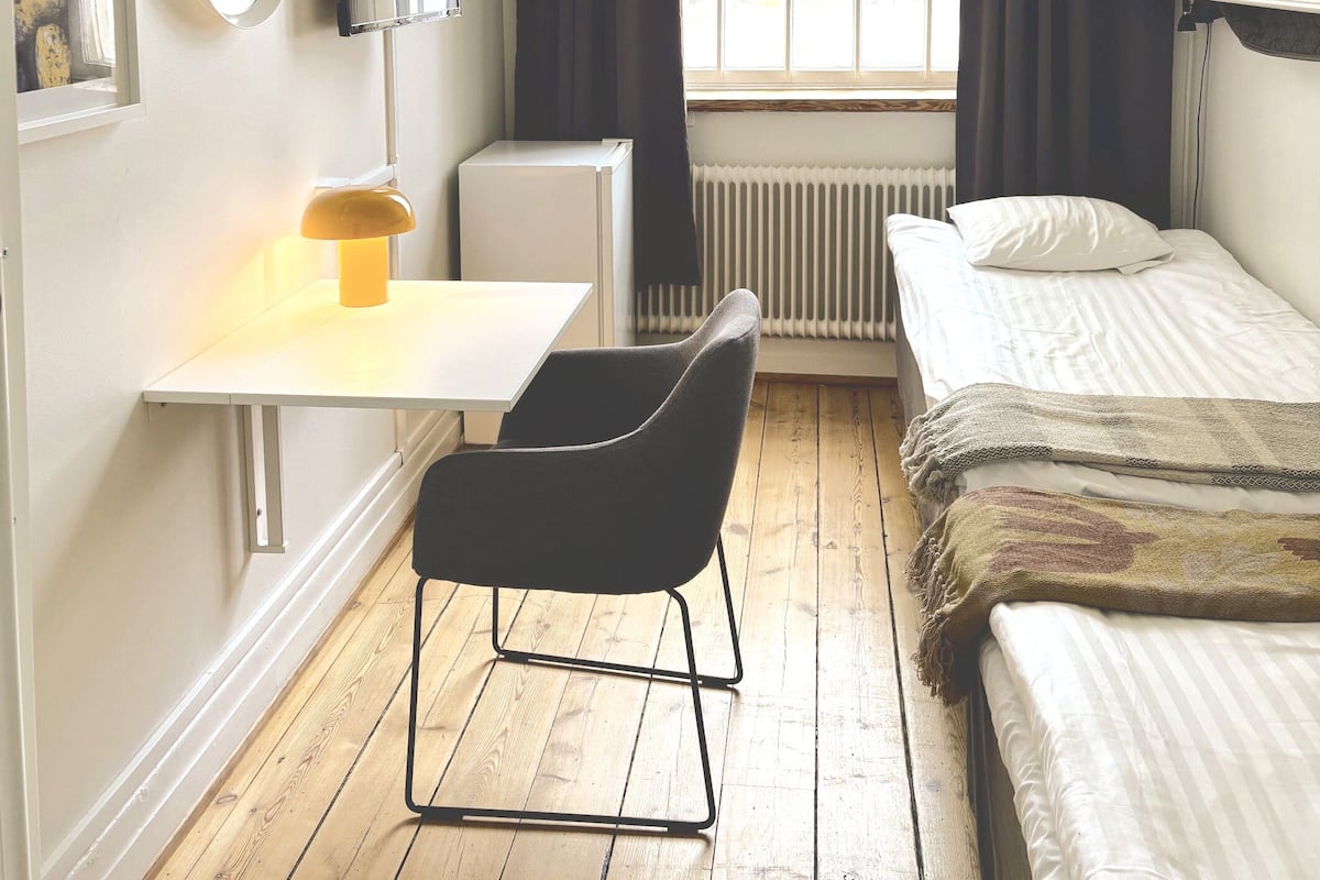 A small, well-lit room with a desk, chair, bed, and a radiator.