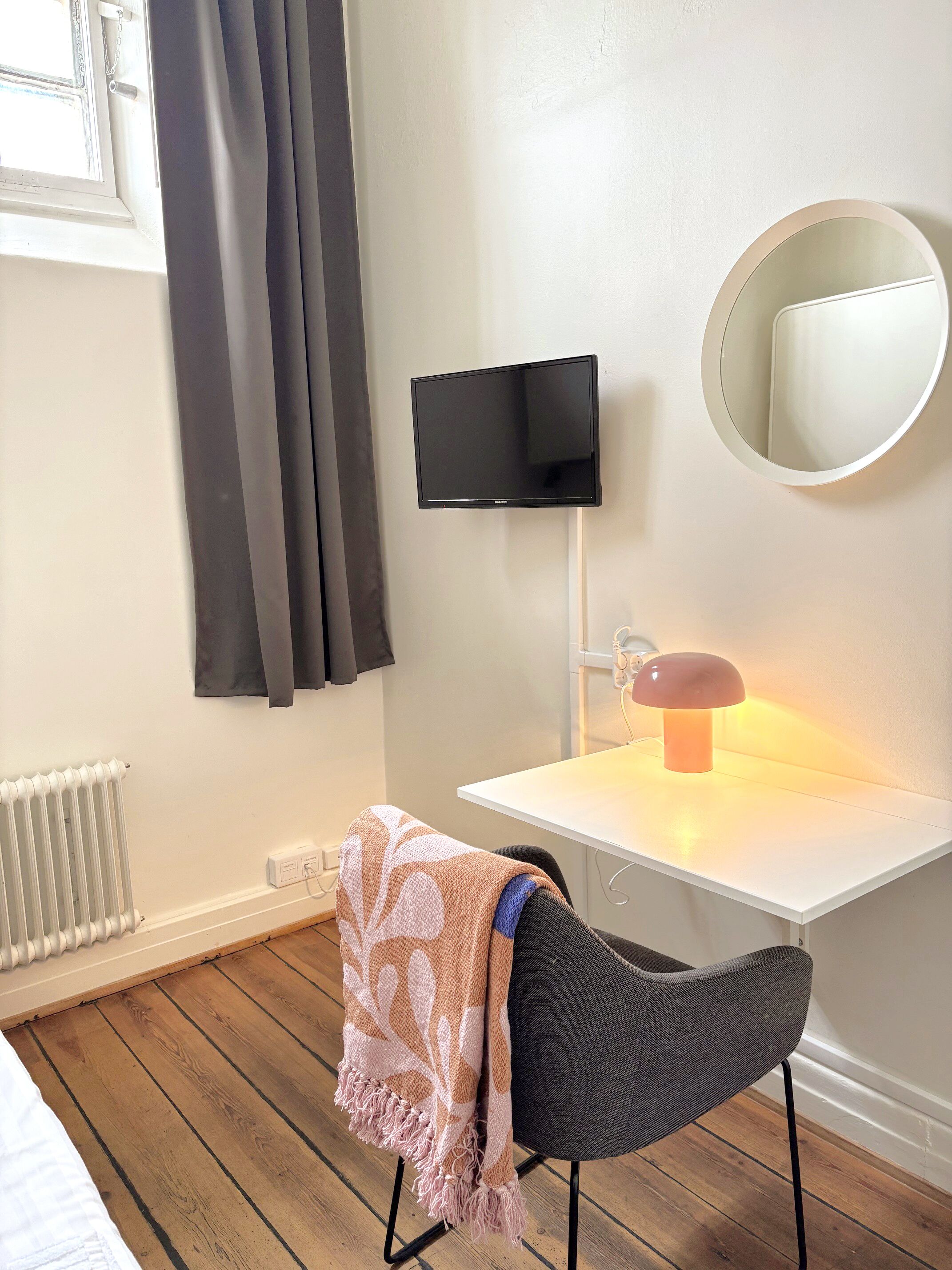 A room with a white desk, a black chair, a round mirror, a flat-screen TV, a heater, and a window with curtains.