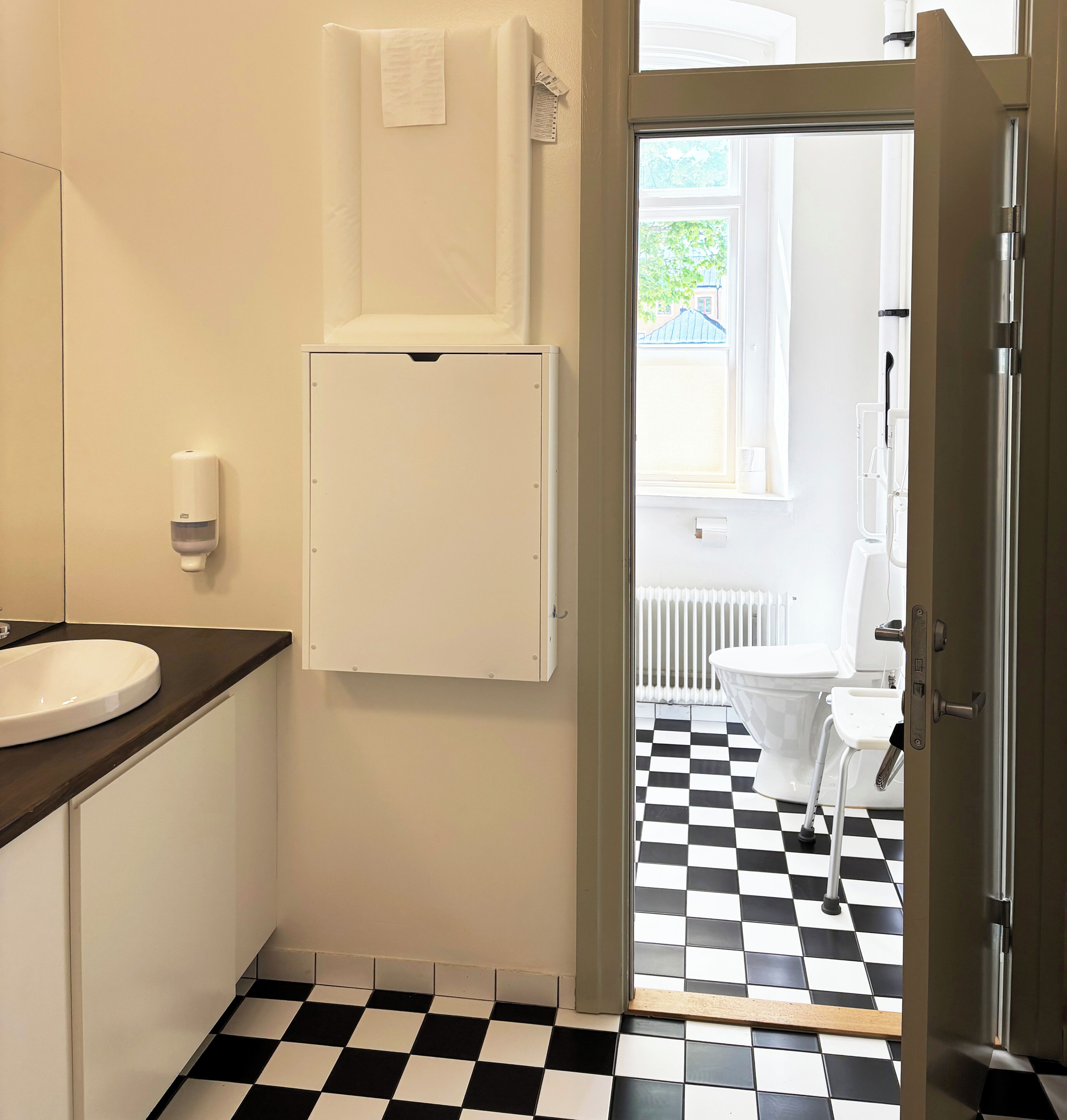 Et badeværelse med en hvid vask, et hvidt skab, gulv i sort og hvid tern og udsigt til en balkon med et hvidt toilet.
