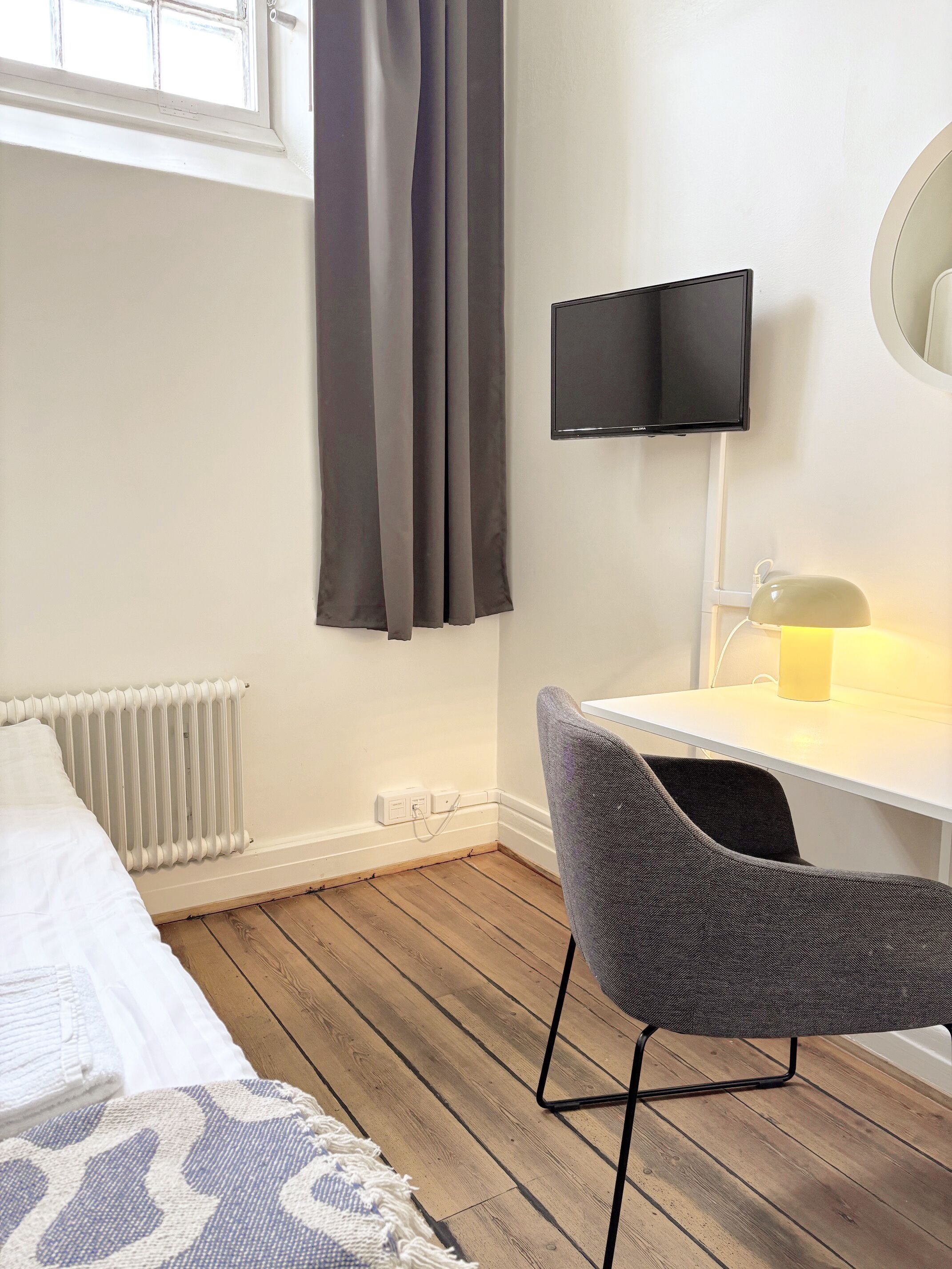 A room with a bed, a desk with a chair, a television, a radiator, and a window with curtains.