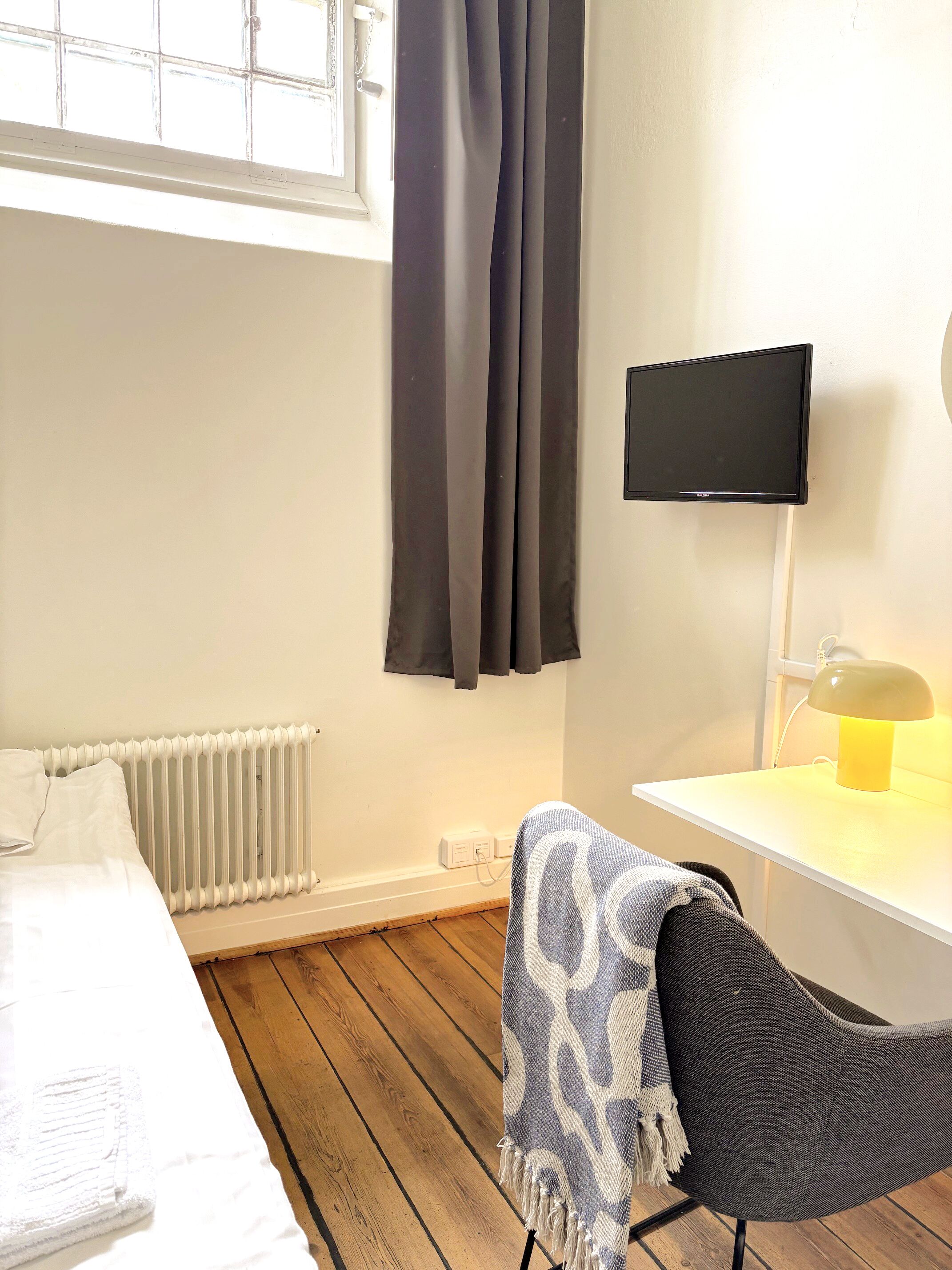 A room with a bed, a desk, a TV, a lamp, and a window with curtains.