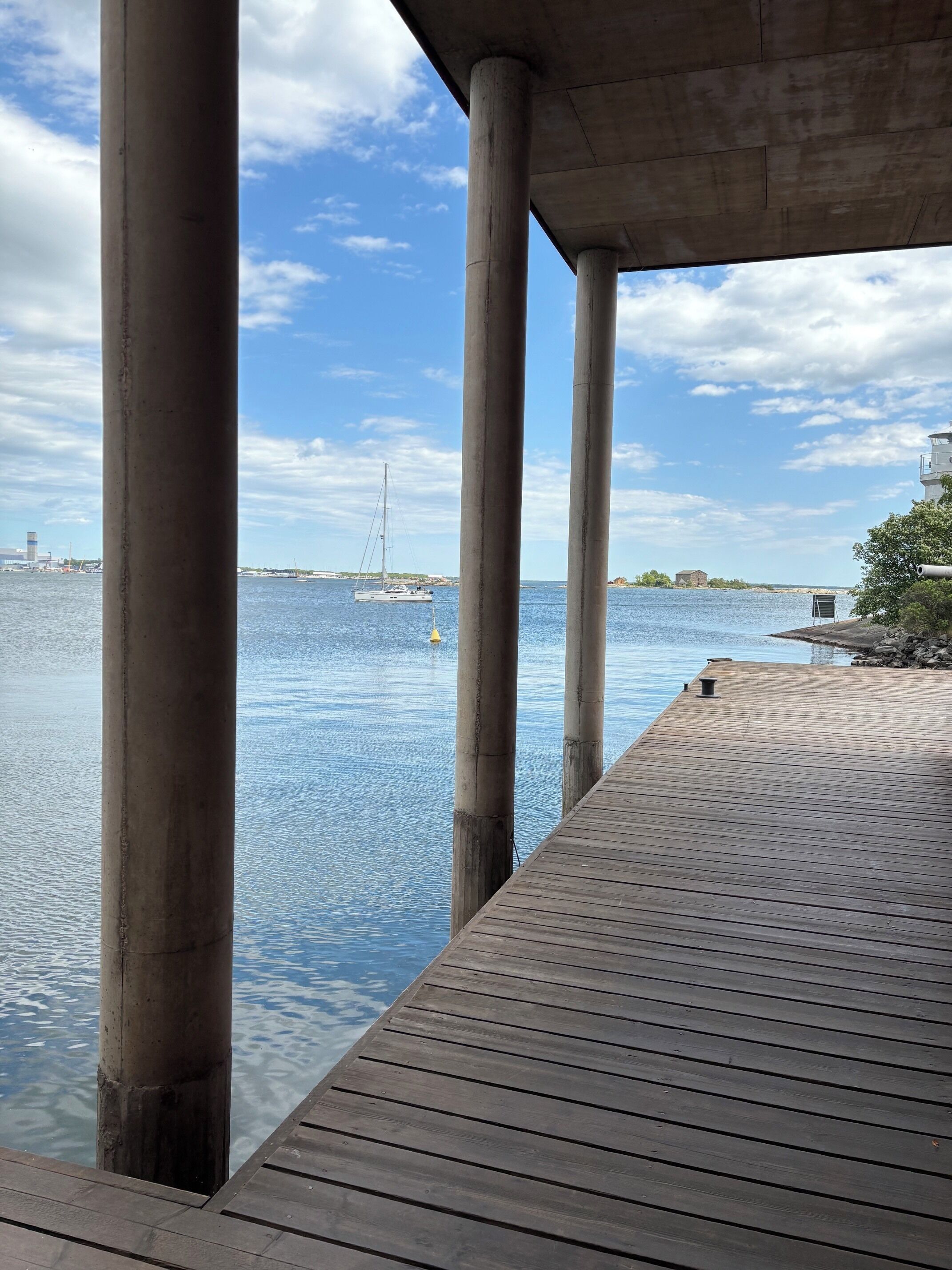 A wooden pier extending into calm water, supported by concrete pillars, with a sailboat in the distance and a clear sky above.