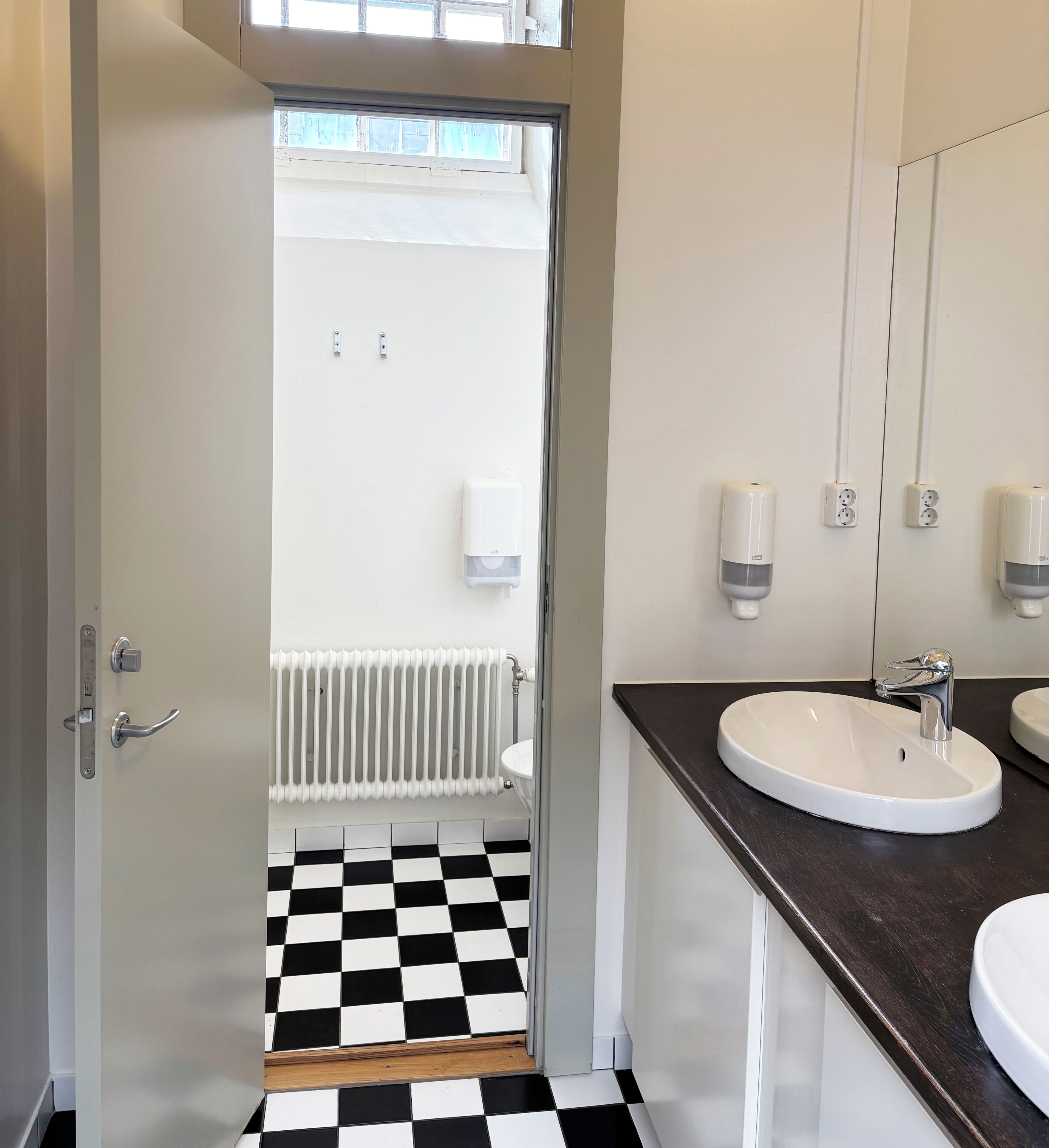 Un baño con suelo a cuadros blanco y negro, un lavabo blanco, un espejo y un radiador.