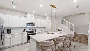 A modern kitchen with white cabinetry, a central island with bar stools, stainless steel appliances, and a staircase leading to an upper level. - 5BR Windsor Cay Home Near Disney, Pool & Games (Clermont)