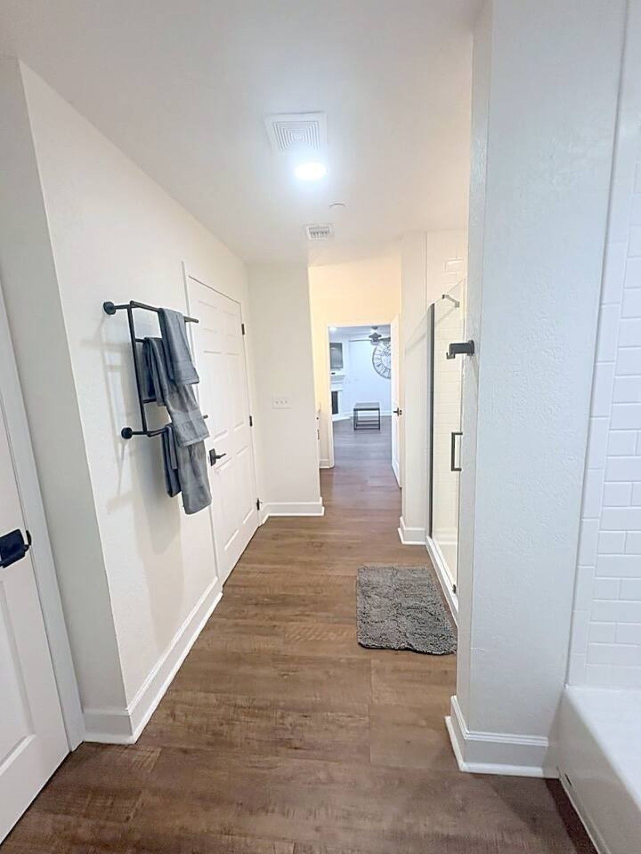 A hallway with white walls, a wooden floor, and a gray rug. There are doors on both sides, and a towel rack on the left.