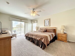 Une chambre à coucher comprenant un lit, un bureau, une chaise, une table de chevet, une lampe, un ventilateur de plafond et une fenêtre donnant sur un beau paysage.