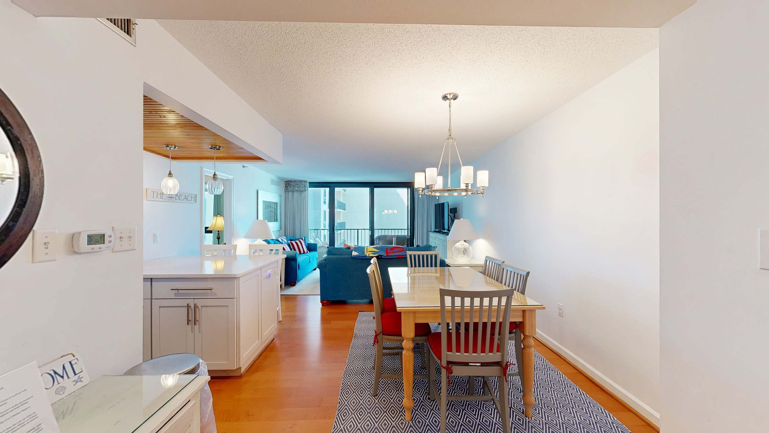 A modern kitchen with a central island, a dining area with a wooden table and chairs, and a living area with a sofa and a view to the outside.