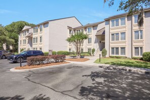 A multi-story residential building with a parking lot, a tree-lined driveway, and a clear blue sky.
