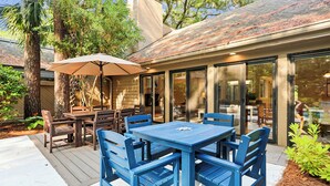 Um pátio externo com mesa e cadeiras, um grande guarda-sol e uma área de refeições com paredes de vidro.