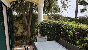 An outdoor patio with a glass-top table and chairs, surrounded by greenery and a yellow building. - Villa lote 10 Paraiso da luz T4 (Luz)
