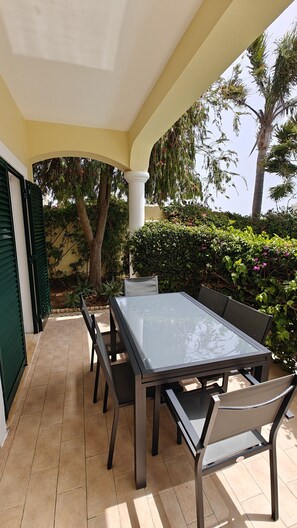 An outdoor patio with a glass-top table and chairs, surrounded by greenery and a yellow building. - Villa lote 10 Paraiso da luz T4 (Luz)