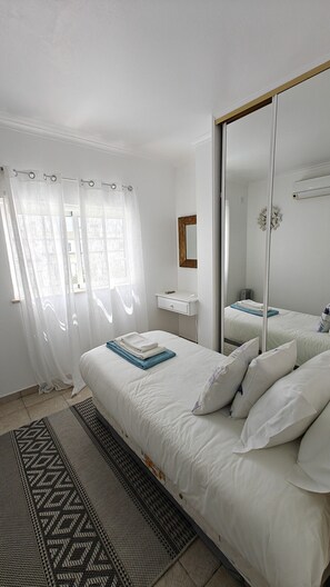 A bedroom with a bed, a bedside table, a mirror, and a window with curtains. - Villa lote 10 Paraiso da luz T4 (Luz)