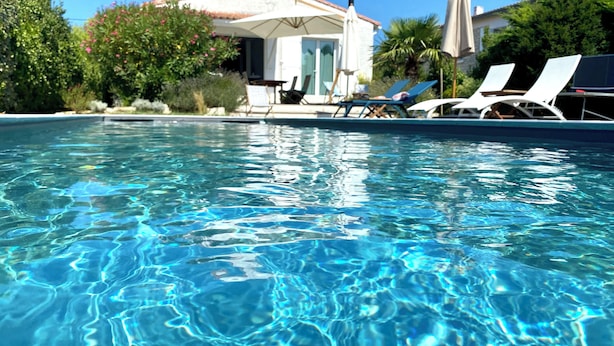 Una piscina cristalina con sillones de descanso y sombrillas, rodeada de vegetación y un edificio con tejado de azulejos.
