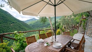 Mesa de madera al aire libre con una sombrilla grande, rodeada de sillas, con vistas a un paisaje montañoso.