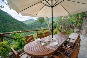 Une table en bois installée à l’extérieur, protégée par un grand parasol et entourée de chaises, d’où l’on peut admirer un paysage montagneux.