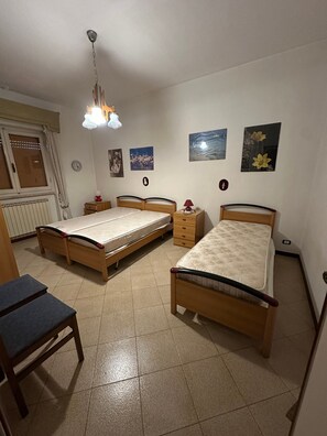 Ein Schlafzimmer mit zwei Einzelbetten, einer hölzernen Bank, einem Nachttisch mit Lampe, einem Heizkörper und einem Fenster mit Rollläden.