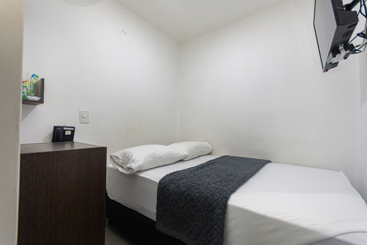 A small, single-bed room with a wooden desk, a white bed, and a dark grey blanket.