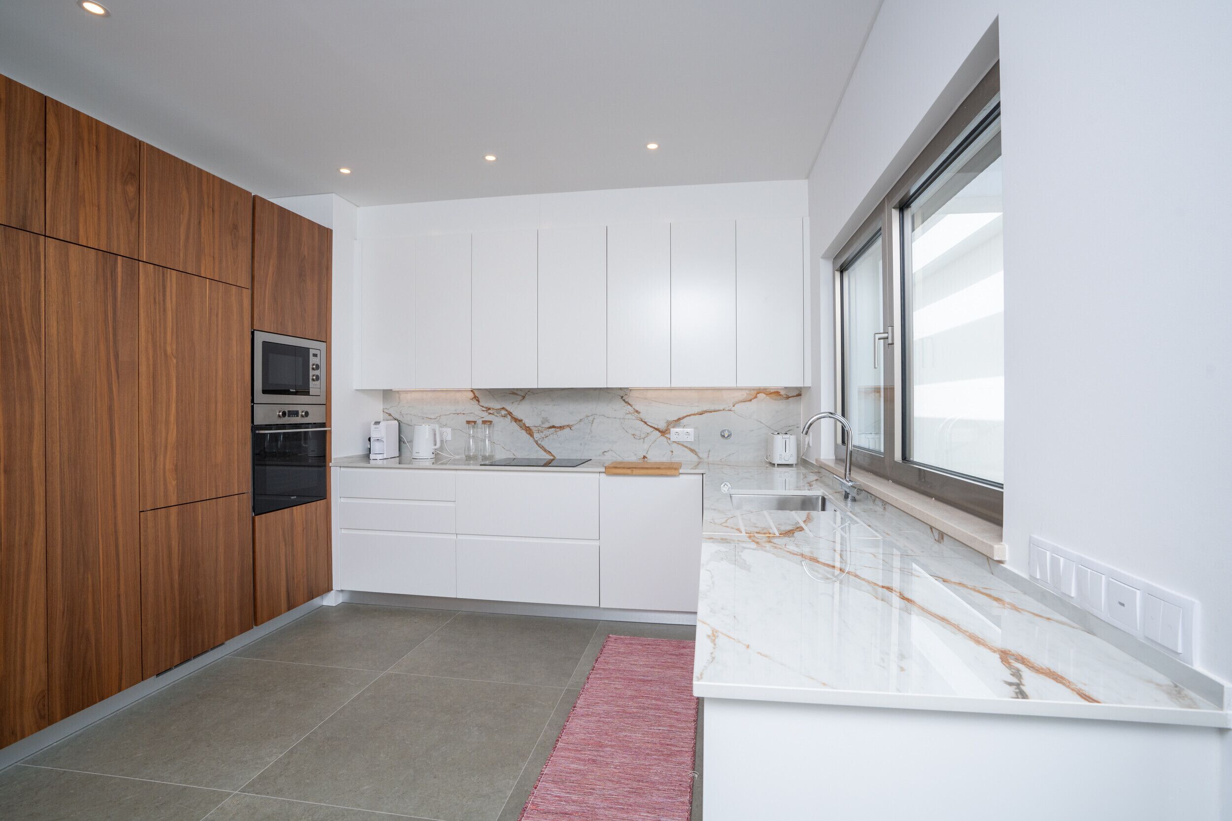 A modern kitchen with wooden cabinets, a built-in oven, white countertops, and a large window.