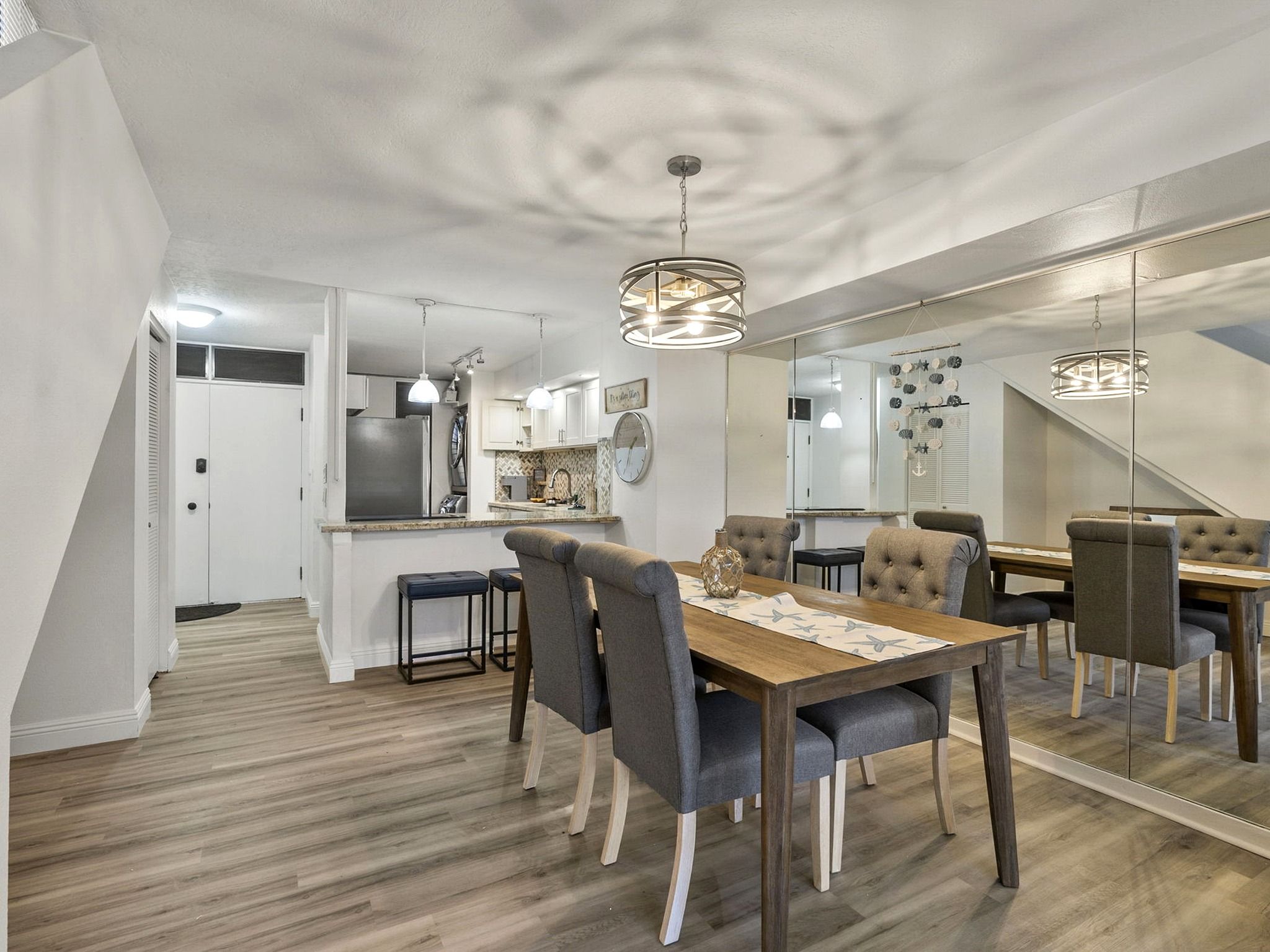 A modern kitchen with a central island, a dining area with a wooden table and upholstered chairs, and a large mirror reflecting the space.