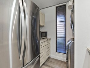 A compact kitchen with a stainless steel refrigerator, a microwave, and a window with blinds. - Stunning Kahana Condo Getaway! (Lahaina)