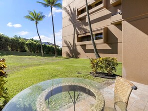 A circular glass table with wicker chairs, a grassy lawn, palm trees, and a building with a beige facade. - Stunning Kahana Condo Getaway! (Lahaina)