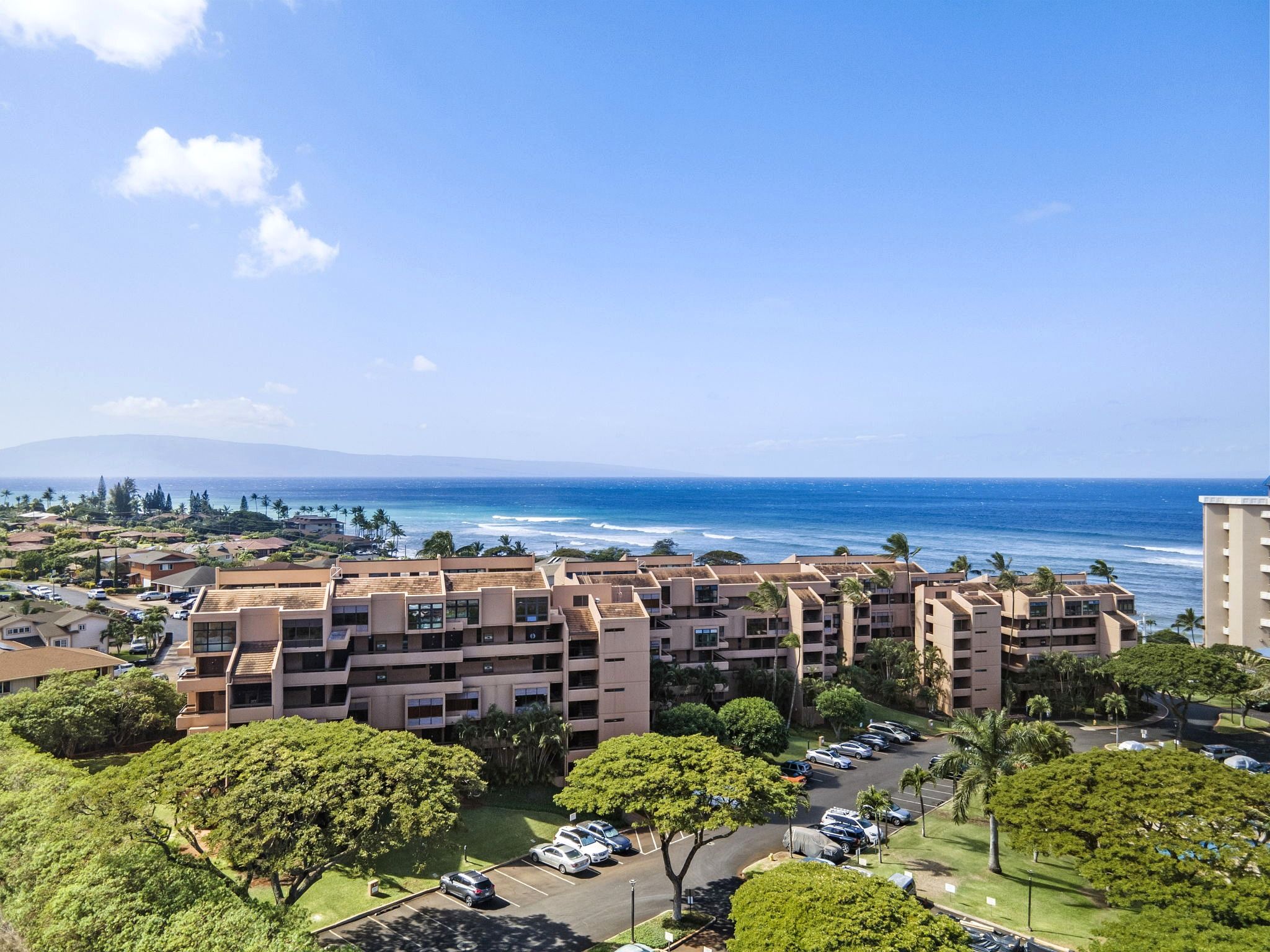 Un hotel de varios pisos con estacionamiento, rodeado de vegetación y vistas a una playa con el océano y una montaña al fondo.