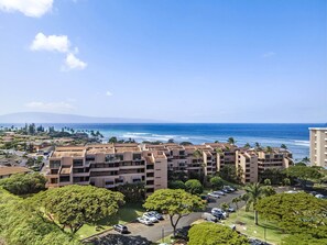 A multi-story hotel with a parking lot, surrounded by greenery and overlooking a beach with the ocean and a mountain in the distance. - Stunning Kahana Condo Getaway! (Lahaina)