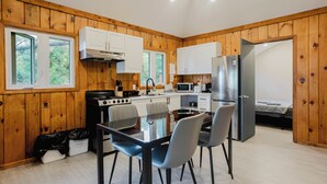A compact kitchen with wooden walls, white cabinets, a dining table with chairs, and a small bed in the corner. - 3-Bedroom Cottage #1 (Nobel)