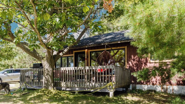 A wooden cabin with a porch, surrounded by trees and a car parked nearby. - 3-Bedroom Cottage #1 (Nobel)