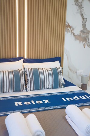 Ein Bett mit gestreiften Kissen, einer blauen Tagesdecke mit dem Aufdruck 'Relax Indulge' und aufgerollten weißen Handtüchern.