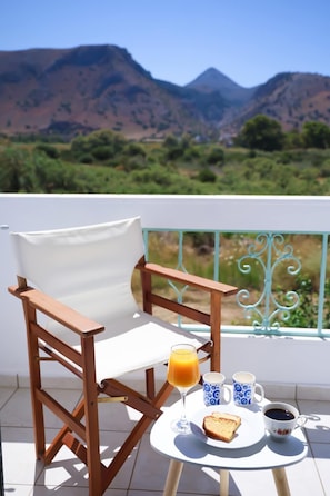 Ein Holzstuhl mit weißem Kissen, ein kleiner runder Tisch mit einem Glas Orangensaft, zwei gemusterte Tassen, eine Kaffeetasse und eine Scheibe Toastbrot.