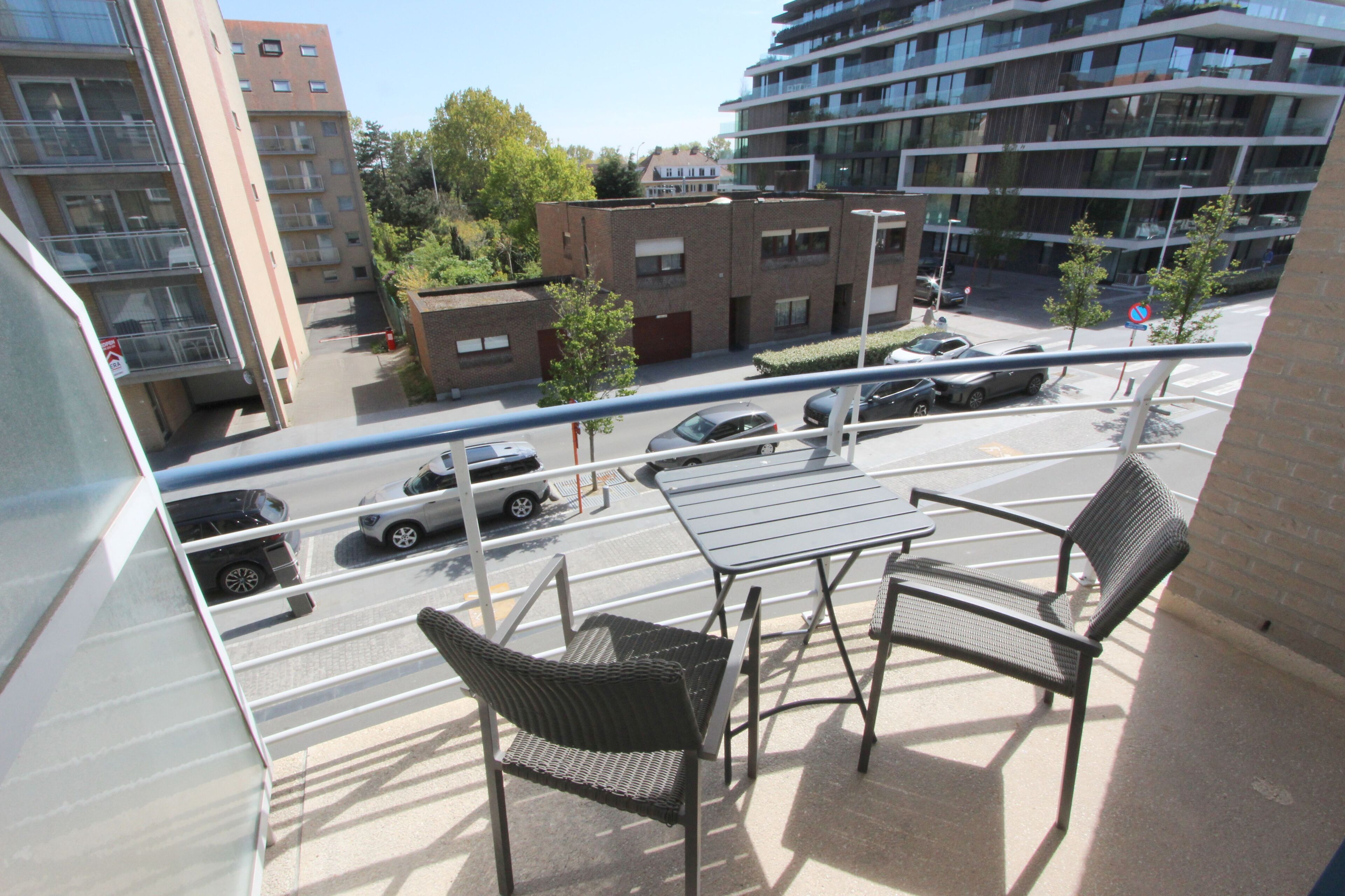 Un balcon avec deux chaises et une table, donnant sur une rue animée par la circulation des voitures et bordée d’immeubles.