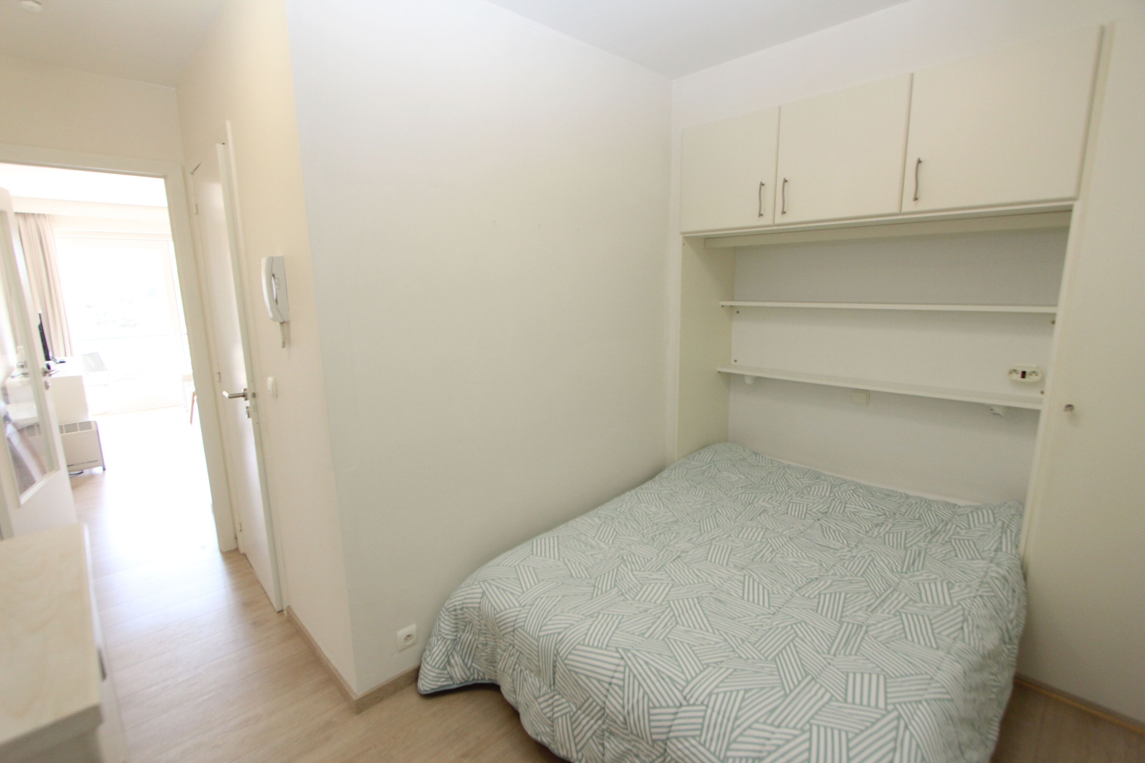 Ein kleines Zimmer mit einem Einzelbett, einer gestreiften Bettdecke, weißen Wänden und eingebauten Regalen über dem Bett.