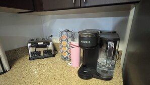 Un taulell de cuina amb un torradora, una cafetera, un got rosa i un recipient de càpsules de cafè.