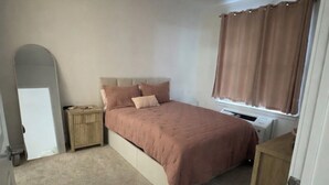 Un dormitori amb un llit, una petita taula de nit, una finestra amb cortines, un radiador i una porta.
