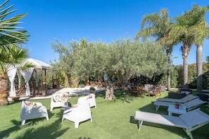 Zona salotto all'aperto con sedie bianche e un tavolo, circondata da alberi di ulivo e un gazebo bianco.
