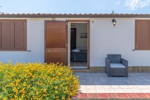 A building with a door open to a room with a bed, a chair outside, and a small garden with yellow flowers. - Room 'Camera Matrimoniale' with Shared Terrace, Wi-Fi and Air Conditioning (Marzamemi)