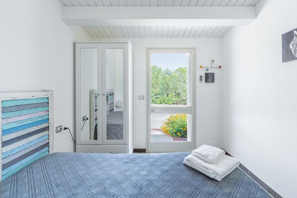 A bedroom with a bed, a door leading to a bathroom, a window with a view of greenery, and a wall-mounted light fixture. - Room 'Camera Matrimoniale' with Shared Terrace, Wi-Fi and Air Conditioning (Marzamemi)
