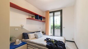 Un dormitorio con una cama, una mesita de noche, un aparador, un ventanal con cortinas y un estante rojo sobre la cama.