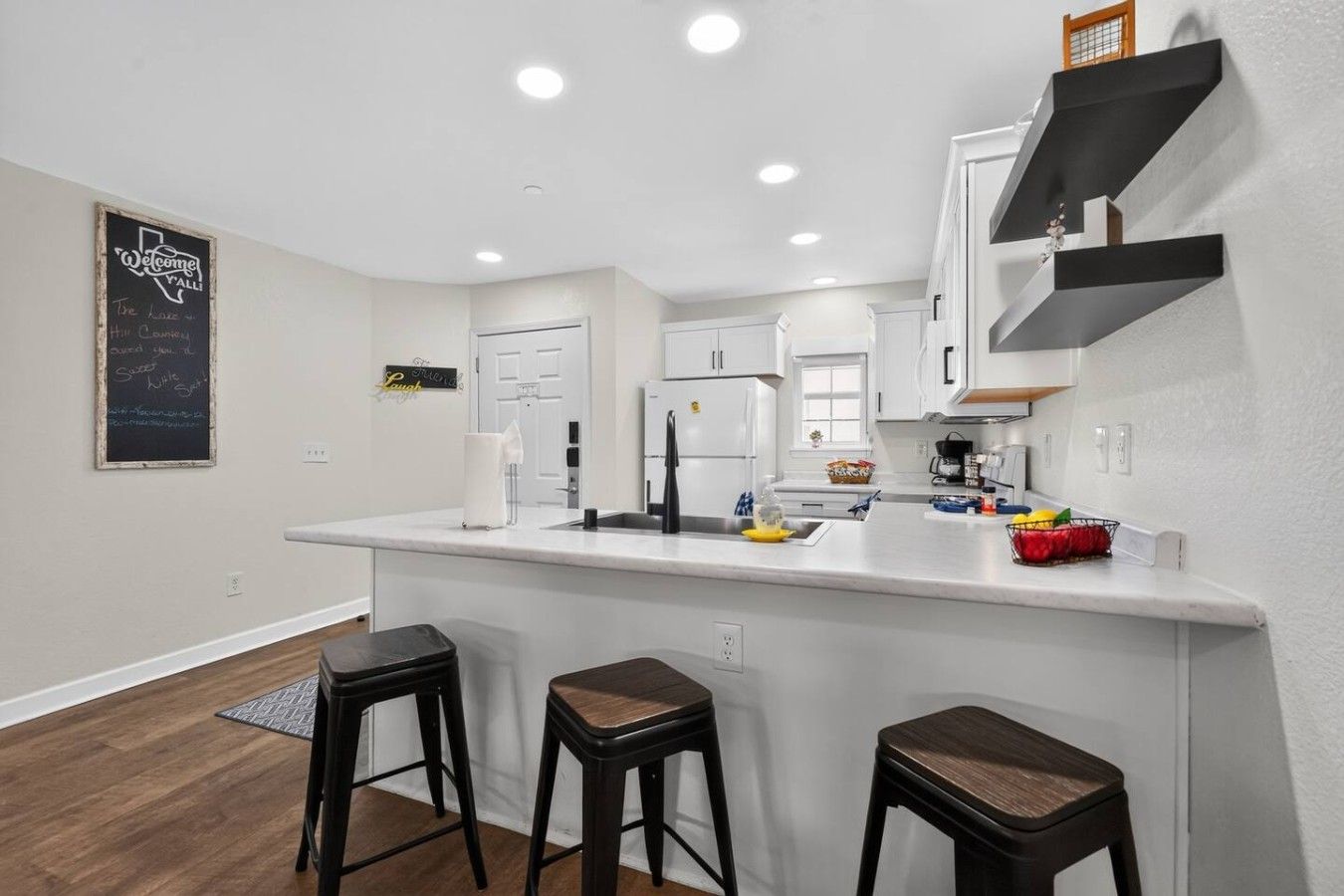 A modern kitchen with a white countertop, black stools, and a chalkboard sign.