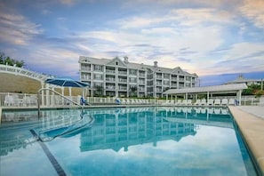 Piscina com espreguiçadeiras, área de estar coberta e edifício com varandas.