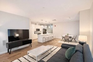 Ein modernes Wohnzimmer mit Fernseher, Sofa, Couchtisch und einer Küche im Hintergrund.