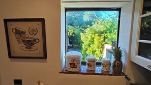 Ein Küchenfenster mit Blick auf Grünflächen, eine gerahmte Kaffeetasse als Kunstwerk und drei Behälter mit Lebensmitteln beschriftet.