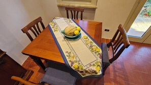 Een eettafel met een tafelkleed met bloemen, een fruitschaal en houten stoelen.