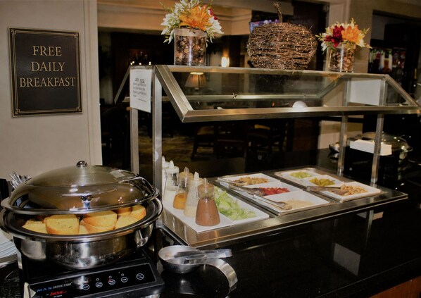 A buffet setup with a sign for free daily breakfast, a variety of food items, condiments, and decorative flowers. - King Suite with Full Kitchen | Free Breakfast (Houston)